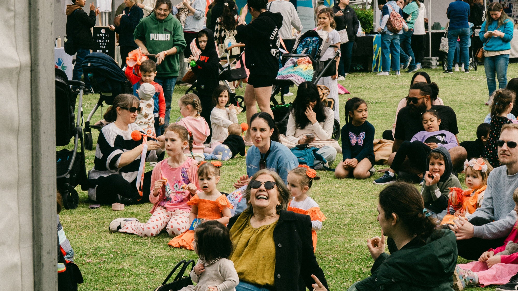 People sitting on grass at an outdoor event with tents in the background